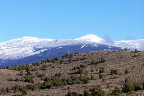 Paisaje montañoso con nieve en las cumbres y vegetación baja salpicada de árboles en los pradillos.
