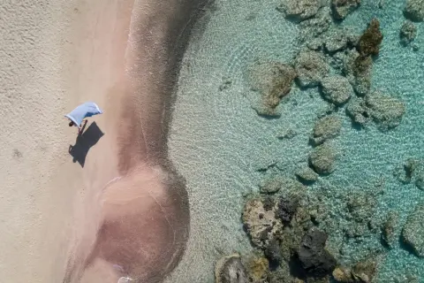 En person med en blå handduk på en rosa strand på Kreta. Klart turkost vatten med stenar.