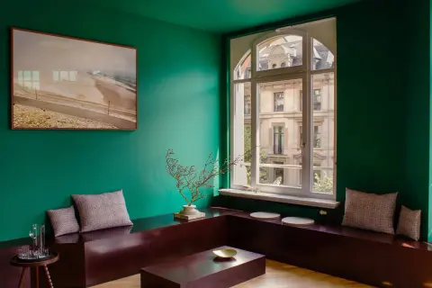 Un salotto con pareti verde scuro, un quadro paesaggistico, sedute in legno scuro e cuscini a quadri. I colori da abbinare al verde scuro includono il marrone del legno e le sfumature neutre dei cuscini.