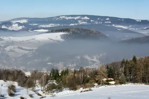 Mgła unosi się nad zimową doliną, ukazując sporą ilość pyłów zawieszonych w powietrzu. W oddali widać ośnieżone wzgórza i lasy.