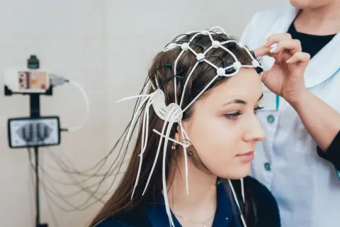 Młoda kobieta z czepkiem EEG na głowie, przygotowywana do badania. W tle aparat do zapisu aktywności mózgu.