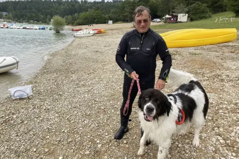 Homme en combinaison de plongée avec son chien noir et blanc sur la plage du lac de Vouglans. Chien autorisé, prêt pour une baignade.