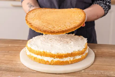 Eine Biskuit-Schicht wird auf eine mit weißer Creme gefüllte Erdbeer-Raffaello-Torte gesetzt.