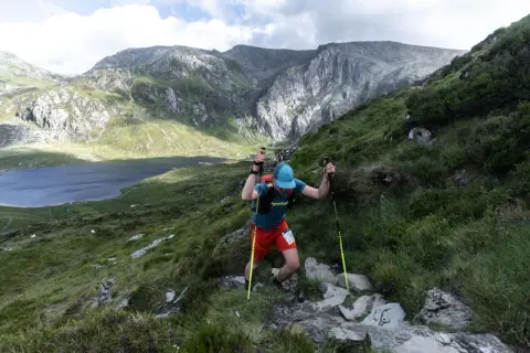 Un coureur grimpe une pente rocheuse, bâtons en main, lors de la kat100 by utmb. Un lac et des montagnes majestueuses en arrière-plan.