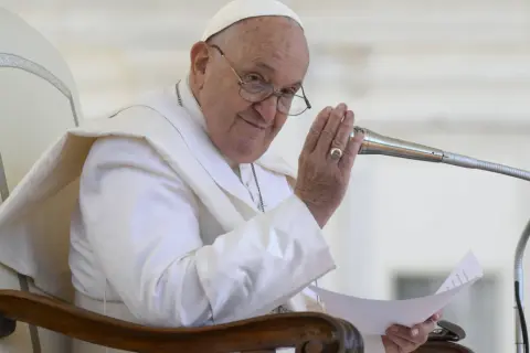 Papież Franciszek w białej szacie siedzi na krześle, trzymając mikrofon i kartkę.