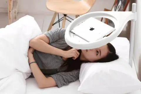 Femme allongée dans un lit, regardant une tablette posée sur un support blanc au-dessus d'elle.