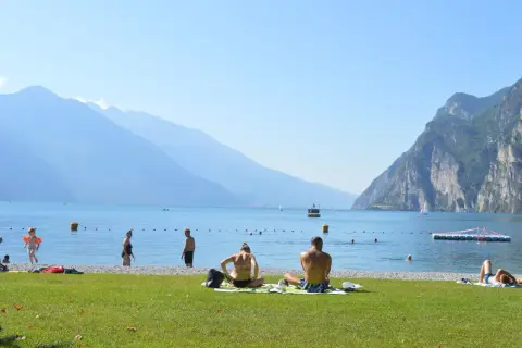 Gente in spiaggia e in acqua, con montagne sullo sfondo, in una giornata di sole sul lago di Garda, forse vicino a Lazise o Bardolino.