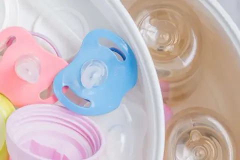 Pink and blue pacifiers sit in a sterilizer with baby bottles. Learn how to sterilize pacifier safely for your baby.