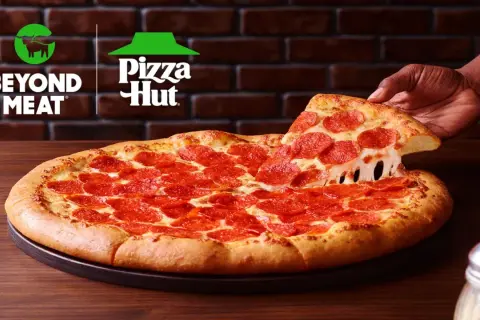 Pizza pepperoni z ciągnącym się serem, podnoszona przez rękę. Logo Beyond Meat i Pizza Hut w tle.
