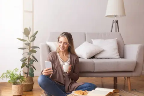 Uśmiechnięta kobieta z telefonem i kawą na podłodze, obok książka i rośliny. Czas na 5 pytań zdrowego myślenia.