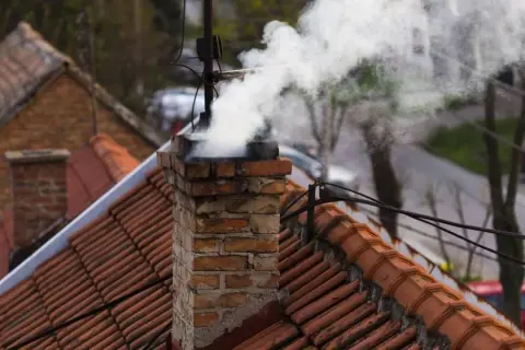Rauch steigt aus einem alten Ziegelschornstein auf. Die Ziegel sind abgenutzt, was auf eine mögliche Schornsteinversottung durch Regen hindeutet.
