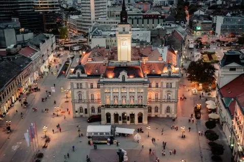 Abendliche Ansicht des Hauptplatzes in Novi Sad, Serbien, mit beleuchteten Gebäuden und Menschen, die spazieren gehen.