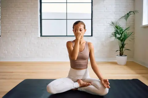 Frau praktiziert Pranayama Atmung im Lotussitz auf einer Yogamatte. Sie hält ihren Finger vor die Nase, um abwechselnd durch die Nasenlöcher zu atmen.