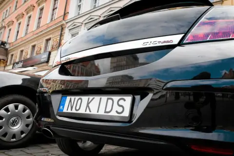 Czarne auto z indywidualnymi tablicami rejestracyjnymi przykłady: "NO KIDS". W tle kamienice.