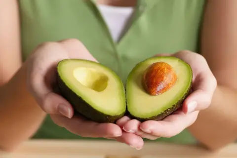 Manos sostienen un aguacate cortado por la mitad, mostrando su pulpa verde y el hueso. Así es qué es el aguacate, una fruta cremosa y nutritiva.