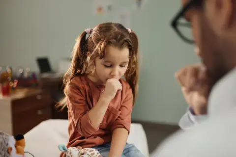 Niña con tos perruna en consulta médica. Un doctor la examina mientras ella se cubre la boca.