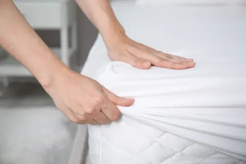 Hands smoothing a white fitted sheet over a mattress. Is it ok to change bed sheets once a month? Probably not for optimal hygiene.