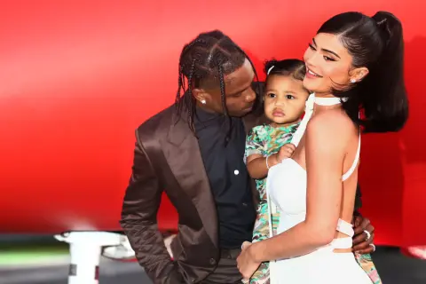 Kylie Jenner z dzieckiem i Travisem Scottem na tle czerwonej ściany.