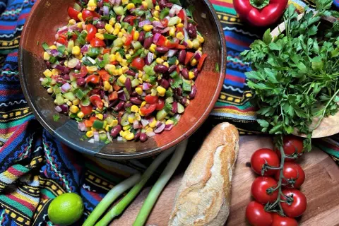 Bunter mexikanischer Salat mit Mais, Bohnen und Gemüse in einer Schüssel, daneben Baguette, Limette und Kräuter.