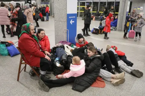 Rodzina z dziećmi, w tym niemowlę, siedzi na podłodze z bagażami, czekając na pomoc. Ich status uchodźcy jest widoczny w ich sytuacji.
