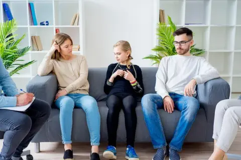 Rodzina podczas terapii systemowej. Terapeuta notuje, rodzice i córka siedzą na kanapie, każdy pogrążony we własnych myślach.