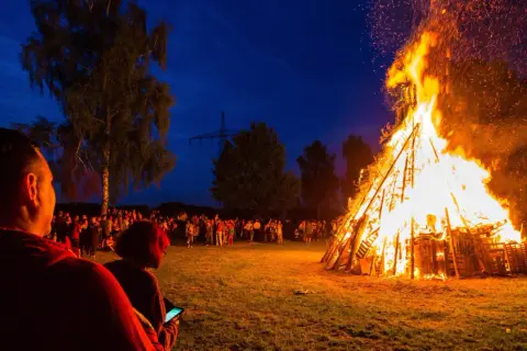 Menschen versammeln sich, um die Sommersonnenwende zu feiern. Ein riesiger Holzstapel brennt lichterloh im Abendhimmel, Funken steigen auf.