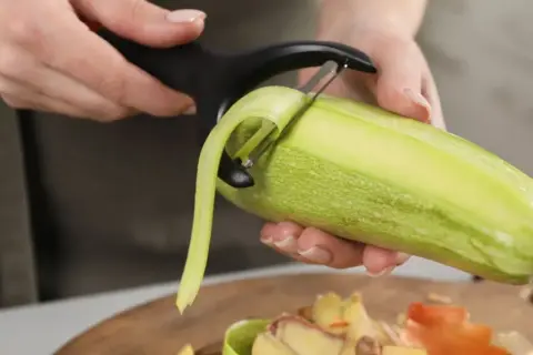 Une personne épluche une courgette. Les épluchures tombent sur une planche à découper, prêtes à être données aux poules.