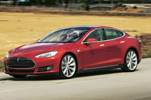 Czerwony Tesla Model S pędzi po drodze, otoczony suchymi, trawiastymi krajobrazami.