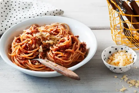 En skål med stuvad spaghetti med köttfärssås och riven ost. En liten skål med mer ost och en korg med bestick står bredvid.