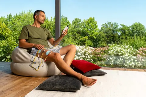 Homme assis, appliquant des électrodes sur sa cuisse, appareil de stimulation en main. Il observe le paysage fleuri, sans effets secondaires apparents du urostim.