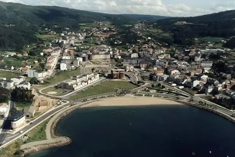 Vista aérea de la playa de Cee, con el mar azul, la arena dorada y la ciudad extendiéndose hacia las colinas verdes.