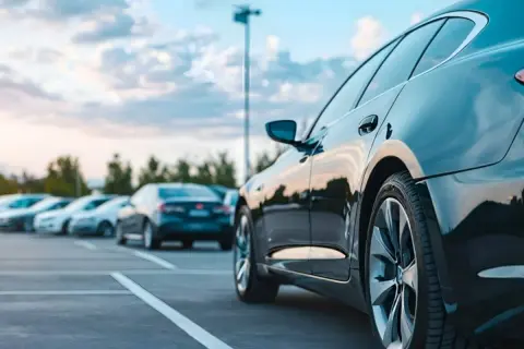 Ciemny lśniący samochód na parkingu, gotowy do rejestracji po. W tle inne auta i niebo z chmurami.