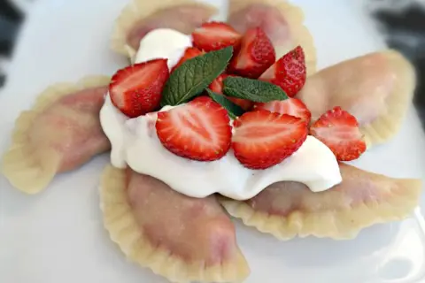 Pyszne pierogi z truskawkami, polane śmietaną i ozdobione świeżymi owocami i miętą. Letnia uczta na talerzu.