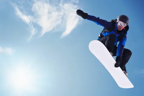 Snowboardzistka w locie na tle błękitnego nieba. Jej deska snowboardowa damska jest biała i lśniąca.