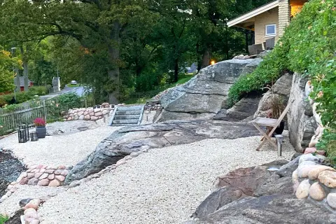 En vacker stenlagd uteplats med grusgångar, stenmurar och trappor som leder upp mot ett hus. En inspiration för en naturlig och robust utomhusmiljö.