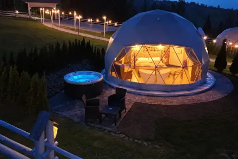 Nocny widok na luksusowy camping w Górach Stołowych. Dwie kopuły z oświetlonym wnętrzem, jacuzzi i meble ogrodowe.