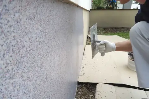 Nakładanie tynku mozaikowego na ścianę budynku. Ręka w białej rękawiczce z kielnią rozprowadza drobne kamyczki.