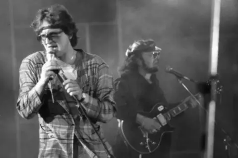 Wokalista z opaską na głowie i w kratę śpiewa do mikrofonu, obok gitarzysta. Widać, że perfect wszystko ma swój czas, nawet na scenie.