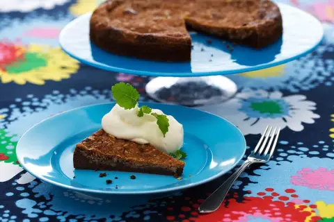 En bit kladdkaka med vispad grädde och mynta på en blå tallrik. Hela kladdkakan syns på en tårtfat i bakgrunden. Ett perfekt kladdkaka recept för fikat.