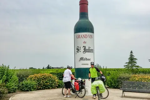 Zwei Radfahrer pausieren vor einer riesigen Weinflasche in Medoc, Frankreich. Weinberge erstrecken sich bis zum Horizont.