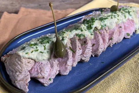 Tranches de vitello tonnato, nappées d'une sauce crémeuse et garnies de câpres et de persil, sur un plat bleu.