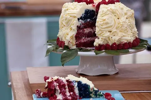 En skivad red velvet cake med vit frosting, hallon och blått inuti. En bit ligger på en blå tallrik.
