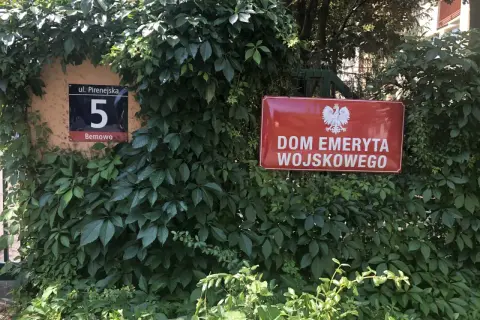 Czerwona tablica z godłem Polski i napisem "DOM EMERYTA WOJSKOWEGO" wśród zieleni. Obok numer domu 5.