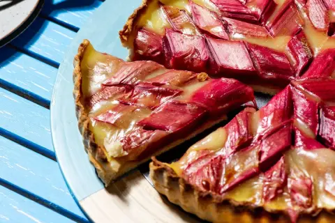 Une tranche de tarte à la rhubarbe, garnie de fruits rouges et d'une crème dorée, repose sur une assiette bleue.