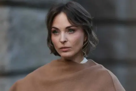 Eine Frau mit Kurzhaarfrisuren und einem leicht molligen Gesicht blickt nachdenklich in die Ferne, eingehüllt in einen braunen Schal.