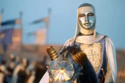 Rycerz w srebrnej masce i zbroi, z herbem Jerozolimy na płaszczu, jedzie konno. To król Baldwin IV.