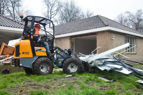 Pomarańczowa koparko-ładowarka z operatorem w kasku i kamizelce usuwa gruz z posesji.