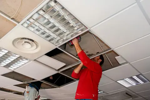 Un hombre con camisa roja instala un panel en **techos registrables**, junto a luminarias y rejillas de ventilación.