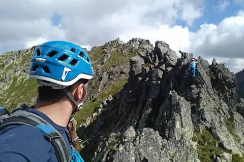 Alpinista w niebieskim kasku patrzy na grań Tatr, gdzie wspina się grupa ludzi.