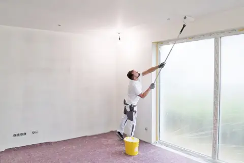 Maler streicht die Decke mit einer Teleskopstange. Ein gelber Farbeimer steht bereit.